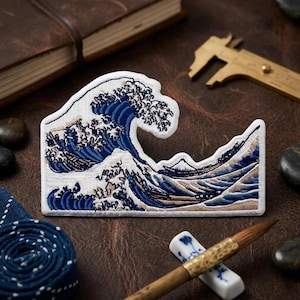May include: An embroidered patch featuring a depiction of a large crashing ocean wave in shades of deep blue, light blue, and white, with a small mountain peak visible in the background, all outlined with a white border on a fabric base.