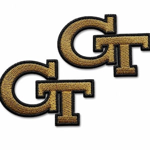 Op de afbeelding: Twee identieke geborduurde patches met de letters GT in een vetgedrukt lettertype. De letters zijn gevuld met metallic goudkleurig garen en omlijnd met een dikke zwarte rand, wat zorgt voor een contrastrijke en textuurrijke uitstraling.