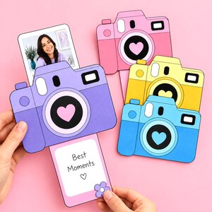 May include: A set of four paper camera cutouts in pink, yellow, blue, and purple, each featuring a heart-shaped lens. One purple camera has a pull-out card displaying the text Best Moments with a heart and flower icon, while another shows a photo.