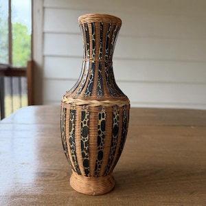 May include: A tall, slender vase constructed from woven light brown wicker and dark brown strips featuring a spotted animal print pattern. The vase has a narrow neck, a rounded body, and a flared base, with a braided wicker band around the center.