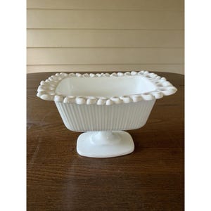May include: A white milk glass rectangular bowl with a pedestal base. The bowl features a ribbed exterior pattern and a decorative looped, scalloped rim. The piece is constructed from opaque white glass with a smooth, glossy finish throughout.