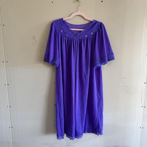 May include: A knee-length purple nightgown featuring short, loose sleeves and a V-neckline. The garment is accented with purple lace trim along the neckline and sleeve edges, and includes small, delicate floral embroidery near the collar area.
