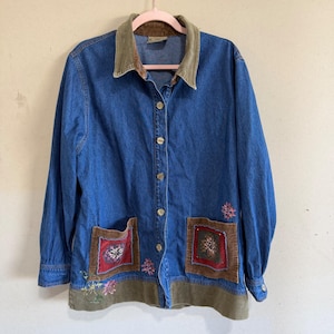 May include: A blue denim long-sleeve shirt featuring a brown corduroy collar and bottom hem. The shirt has a button-down front, two front patch pockets with brown corduroy trim, and embroidered floral details in pink, green, and white colors.