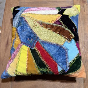 May include: A square throw pillow featuring a patchwork design of various velvet and fabric scraps in shades of yellow, blue, red, green, and black. The pieces are joined together with visible decorative embroidery stitching in contrasting colors.