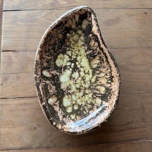May include: An asymmetrical ceramic dish with a teardrop shape, featuring a mottled glaze in shades of dark brown, cream, and soft pink. The interior surface displays a textured, crystalline pattern with dark brown veins radiating outward.