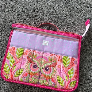 May include: A rectangular fabric carrying case with a zipper closure and a top handle. The front features a pink panel with a colorful owl and leaf pattern, a lavender section with small green stars, and a label reading Jen Laping Designs.