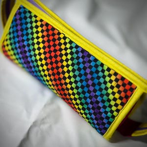 May include: A rectangular, quilted pencil case with a vibrant rainbow checkerboard pattern. The case is trimmed with bright yellow fabric and features a red strap. The interior is lined with purple fabric.