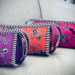 May include: Three quilted cosmetic bags in vibrant colors: purple, orange, and pink. Each bag features a floral pattern with a bat silhouette, black and white striped handles and trim. The bags are arranged on a neutral surface.
