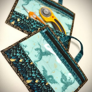 May include: Two rectangular zippered pouches with handles, featuring a clear vinyl window displaying a light blue fabric with dark teal dragon illustrations and gold stars. The lower section has a dark teal and black dragon scale pattern.