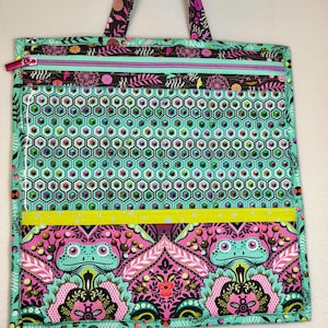 May include: A square fabric tote bag featuring a top handle and a zippered closure. The design includes a clear plastic section over a teal and black geometric honeycomb pattern, a bright yellow horizontal stripe, and a bottom panel with frogs.