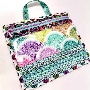 May include: A rectangular zippered pouch with a top handle, featuring a clear front panel. The design includes a vibrant pattern of purple, green, and teal fan-shaped motifs, a lower section with a teal honeycomb print, and a bright teal zipper.