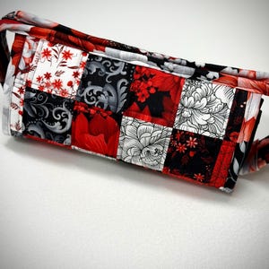 May include: A quilted bag with a patchwork design featuring red, black, white, and gray floral patterns. The bag's fabric is divided into squares, each showcasing a different floral print. The bag has a handle and is set against a white background.