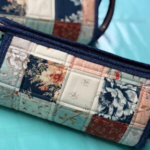May include: Quilted patchwork clutch with a navy blue trim. The fabric features floral patterns in shades of blue, pink, and cream. The clutch has a rectangular shape and a quilted texture, with a matching handle.