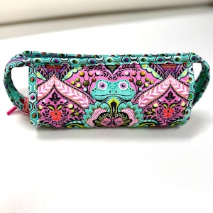 May include: A rectangular fabric pencil case featuring a vibrant pattern of a stylized light blue frog centered among pink, black, and teal floral and geometric motifs. The case has a teal hexagonal border and a pink zipper pull on the side.