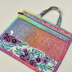 May include: A clear, square tote bag with a colorful, patterned design. The bag features a zipper closure and a handle. The bottom of the bag has a floral pattern with purple flowers and green leaves.