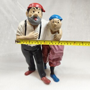 Op de afbeelding: Een geschilderd beeldje van twee figuren die samen staan, &eacute;&eacute;n met een rode pet en grijze baard, de andere met een blauwe pet. Een geel meetlint ligt ervoor en toont een breedte van ongeveer 28 cm over de figuren heen.
