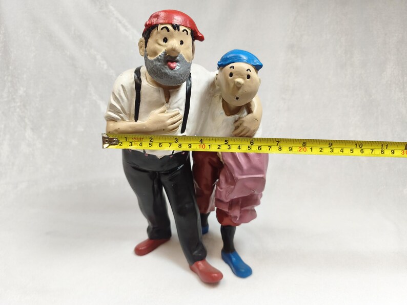 Op de afbeelding: Een geschilderd beeldje van twee figuren die samen staan, &eacute;&eacute;n met een rode pet en grijze baard, de andere met een blauwe pet. Een geel meetlint ligt ervoor en toont een breedte van ongeveer 28 cm over de figuren heen.