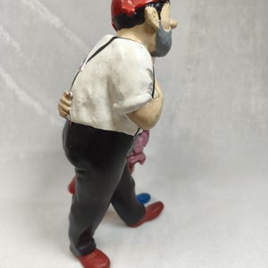 Op de afbeelding: Een beeldje van een persoon met een rode muts, een wit overhemd met zwarte bretels en een zwarte broek. Het figuur heeft een grijze baard en is afgebeeld in zijaanzicht, met &eacute;&eacute;n hand achter de rug en rode schoenen aan de voeten.