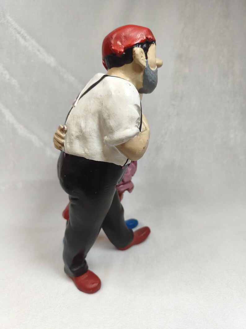 Op de afbeelding: Een beeldje van een persoon met een rode muts, een wit overhemd met zwarte bretels en een zwarte broek. Het figuur heeft een grijze baard en is afgebeeld in zijaanzicht, met &eacute;&eacute;n hand achter de rug en rode schoenen aan de voeten.