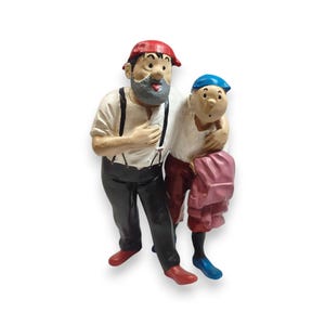Op de afbeelding: Een geschilderd beeldje van twee figuren die samen staan. De langere figuur heeft een grijze baard, een rode pet, een wit overhemd met zwarte bretels en een zwarte broek. De kleinere figuur draagt een blauwe pet en een wit overhemd.