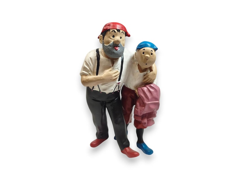 Op de afbeelding: Een geschilderd beeldje van twee figuren die samen staan. De langere figuur heeft een grijze baard, een rode pet, een wit overhemd met zwarte bretels en een zwarte broek. De kleinere figuur draagt een blauwe pet en een wit overhemd.