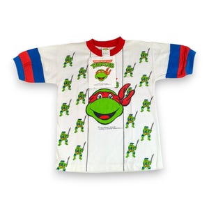 Pode incluir: Uma t-shirt branca de manga curta com um padrão repetitivo de personagens das Teenage Mutant Ninja Turtles, um grande gráfico central de um rosto de tartaruga com máscara vermelha, gola vermelha e punhos às riscas azuis e vermelhas.