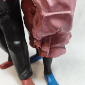 Op de afbeelding: Een beeldje van twee figuren die samen staan. E&eacute;n figuur draagt een zwarte broek met rode schoenen, terwijl de andere een roze kledingstuk met blauwe schoenen draagt. De figuren zijn gemaakt van een glad, beschilderd materiaal.
