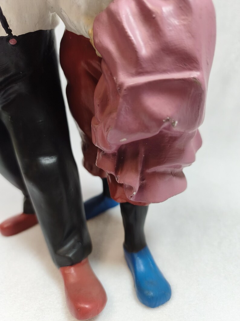 Op de afbeelding: Een beeldje van twee figuren die samen staan. E&eacute;n figuur draagt een zwarte broek met rode schoenen, terwijl de andere een roze kledingstuk met blauwe schoenen draagt. De figuren zijn gemaakt van een glad, beschilderd materiaal.