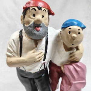 Op de afbeelding: Een geschilderd beeldje van twee figuren die naast elkaar staan. De langere figuur draagt een rode pet, een grijze baard, een wit overhemd, zwarte bretels en een zwarte broek. De kortere figuur draagt een blauwe pet en een donkerrode broek.