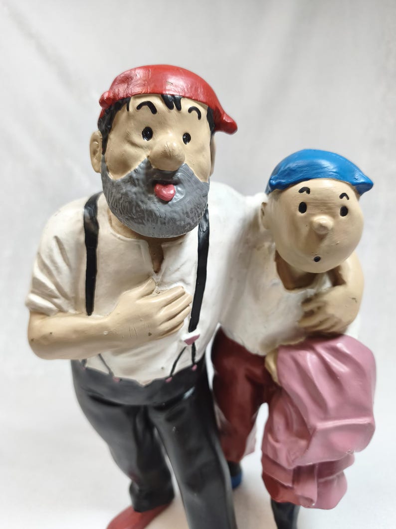 Op de afbeelding: Een geschilderd beeldje van twee figuren die naast elkaar staan. De langere figuur draagt een rode pet, een grijze baard, een wit overhemd, zwarte bretels en een zwarte broek. De kortere figuur draagt een blauwe pet en een donkerrode broek.