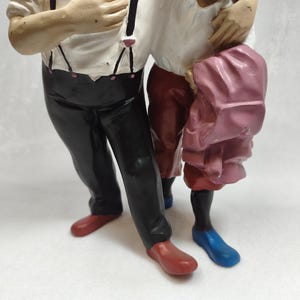 Op de afbeelding: Een beeldje met twee figuren die naast elkaar staan. E&eacute;n figuur draagt een wit overhemd met zwarte bretels, een zwarte broek en rode schoenen. Het andere figuur draagt een wit overhemd, een donkerrode broek, blauwe schoenen en houdt een roze kledingstuk vast.