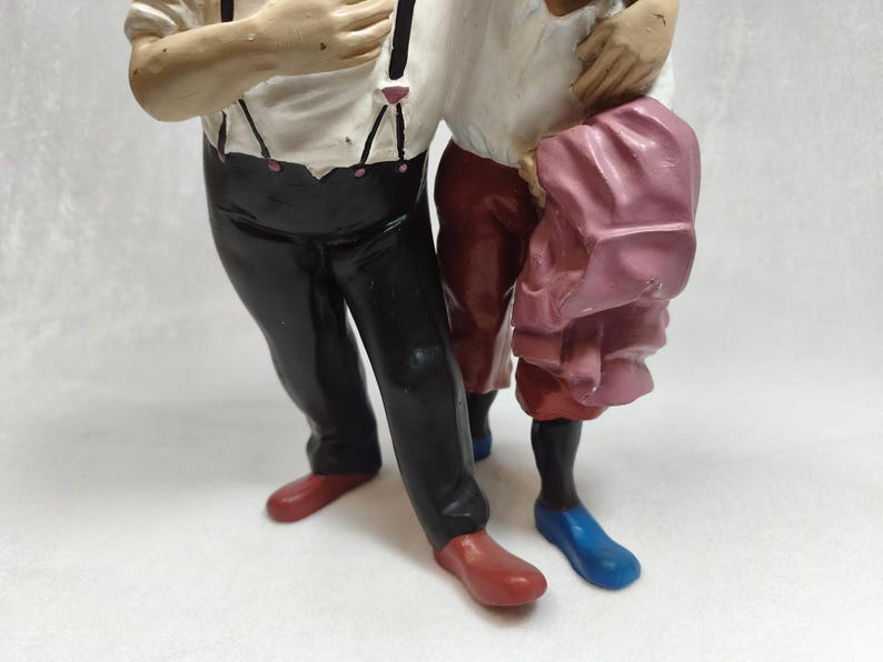 Op de afbeelding: Een beeldje met twee figuren die naast elkaar staan. E&eacute;n figuur draagt een wit overhemd met zwarte bretels, een zwarte broek en rode schoenen. Het andere figuur draagt een wit overhemd, een donkerrode broek, blauwe schoenen en houdt een roze kledingstuk vast.