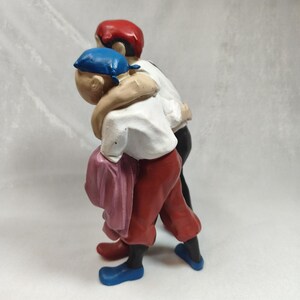 Op de afbeelding: Een beeldje van twee figuren die dicht bij elkaar staan met hun armen om elkaar heen. E&eacute;n figuur draagt een blauwe hoofdbedekking, een wit overhemd, een rode broek en blauwe schoenen. De andere figuur draagt een rode hoofdbedekking, een wit overhemd en een zwarte broek.