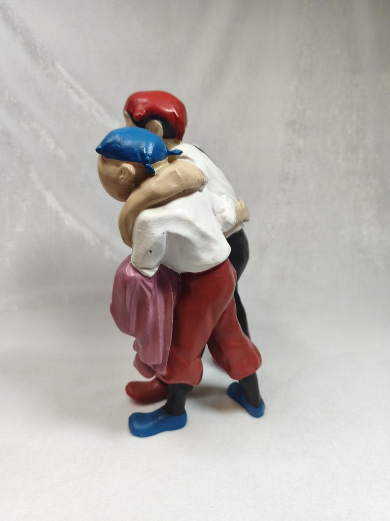 Op de afbeelding: Een beeldje van twee figuren die dicht bij elkaar staan met hun armen om elkaar heen. E&eacute;n figuur draagt een blauwe hoofdbedekking, een wit overhemd, een rode broek en blauwe schoenen. De andere figuur draagt een rode hoofdbedekking, een wit overhemd en een zwarte broek.
