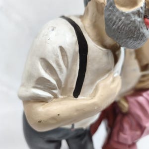 Op de afbeelding: Een geschilderd beeldje van een persoon met een grijze baard, gekleed in een wit shirt met korte mouwen, zwarte bretels en een donkergrijze broek. Het figuurtje draagt een rode hoed en staat naast een ander figuur in roze kleding.