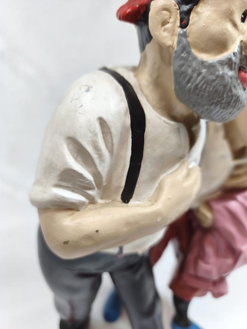 Op de afbeelding: Een geschilderd beeldje van een persoon met een grijze baard, gekleed in een wit shirt met korte mouwen, zwarte bretels en een donkergrijze broek. Het figuurtje draagt een rode hoed en staat naast een ander figuur in roze kleding.