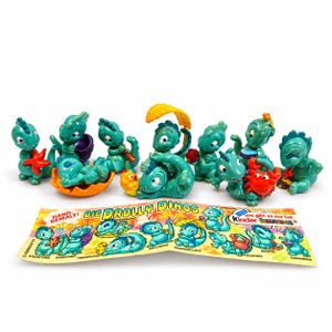 May include: A collection of eight small teal dinosaur figurines, each with unique poses and accessories, accompanied by a paper display insert featuring the text DIE DROLLY DINOS and Kinder Überraschung, showcasing the various character designs.