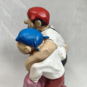 Op de afbeelding: Een beschilderd beeldje van twee figuren in een omhelzing. E&eacute;n figuur draagt een felblauwe hoofdbedekking en een wit overhemd, terwijl de andere figuur een rode hoofdbedekking, een wit overhemd en een donkere broek draagt, met een roze doek eronder.