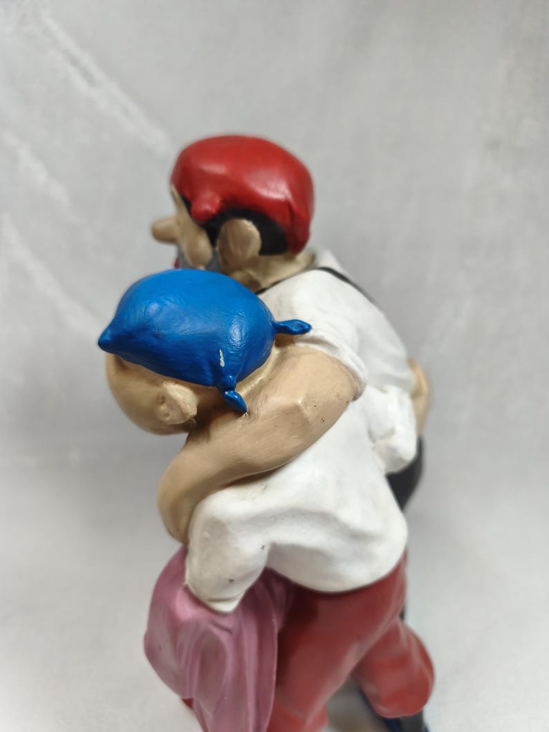 Op de afbeelding: Een beschilderd beeldje van twee figuren in een omhelzing. E&eacute;n figuur draagt een felblauwe hoofdbedekking en een wit overhemd, terwijl de andere figuur een rode hoofdbedekking, een wit overhemd en een donkere broek draagt, met een roze doek eronder.