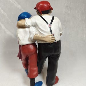 Op de afbeelding: Een beeldje van twee figuren die zij aan zij staan met hun armen om elkaar heen. Het grotere figuur draagt een rode pet, een wit overhemd met zwarte bretels en een zwarte broek. Het kleinere figuur draagt een blauwe pet, wit overhemd en rode broek.