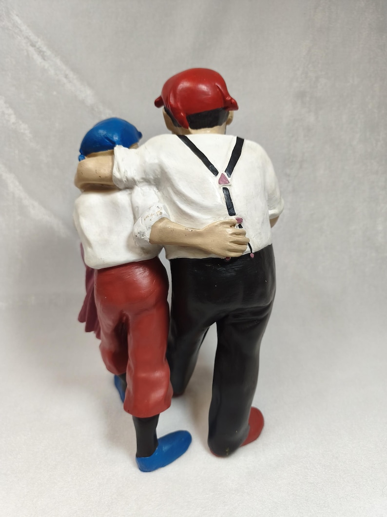 Op de afbeelding: Een beeldje van twee figuren die zij aan zij staan met hun armen om elkaar heen. Het grotere figuur draagt een rode pet, een wit overhemd met zwarte bretels en een zwarte broek. Het kleinere figuur draagt een blauwe pet, wit overhemd en rode broek.