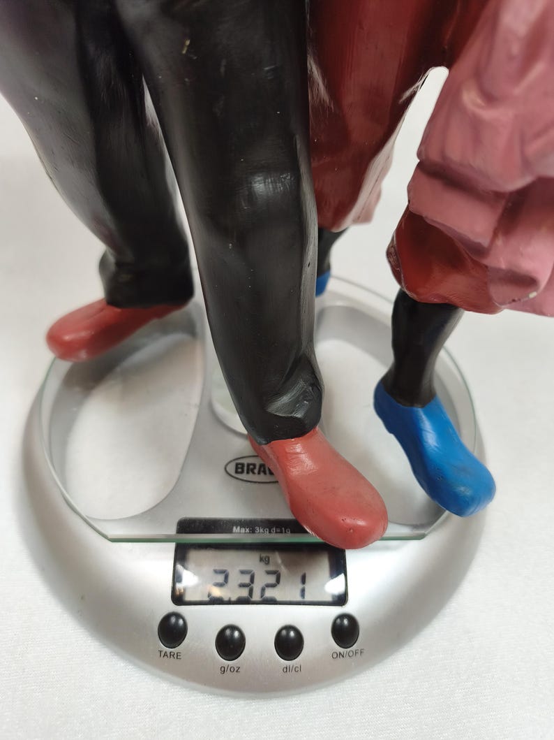 Op de afbeelding: Een veelkleurig beeldje met een zwarte broek, rode schoenen en blauwe schoenen op een zilveren digitale keukenweegschaal. Het display van de weegschaal toont een gewicht van 2,321 kg, met de knoppen TARE, g/oz, dl/cl en ON/OFF.