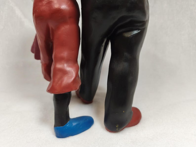 Op de afbeelding: Een beeldje dat de onderlichamen van twee figuren toont. E&eacute;n figuur draagt een rode broek en een blauwe schoen, terwijl de andere figuur een zwarte broek en een rode schoen draagt. De figuren staan naast elkaar met een gladde, geverfde afwerking.