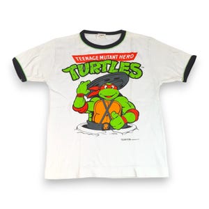 May include: A white short-sleeve t-shirt with black trim on the collar and sleeves. The front features a graphic of a green Teenage Mutant Hero Turtles character wearing a red mask and orange chest plate, emerging from a manhole cover.