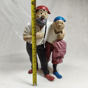 Op de afbeelding: Een geschilderd beeldje van twee figuren die samen staan, met een geel meetlint dat een hoogte van ongeveer 30 cm aangeeft. De grotere figuur draagt een rode pet, wit overhemd, zwarte broek en rode schoenen. De kleinere figuur draagt een blauwe pet, wit overhemd, rode broek en blauwe schoenen.