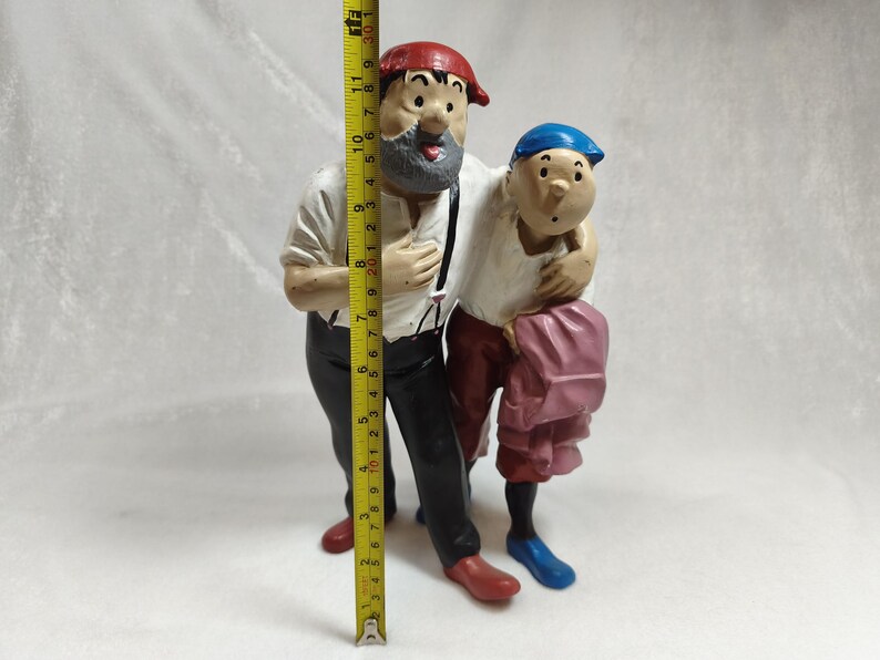 Op de afbeelding: Een geschilderd beeldje van twee figuren die samen staan, met een geel meetlint dat een hoogte van ongeveer 30 cm aangeeft. De grotere figuur draagt een rode pet, wit overhemd, zwarte broek en rode schoenen. De kleinere figuur draagt een blauwe pet, wit overhemd, rode broek en blauwe schoenen.