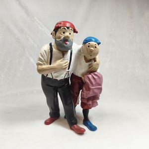 Op de afbeelding: Een geschilderd beeldje van twee figuren die samen staan. Het langere figuur heeft een grijze baard, een rode pet, een wit overhemd met zwarte bretels, een zwarte broek en rode schoenen. Het kleinere figuur draagt een blauwe pet en roze broek.