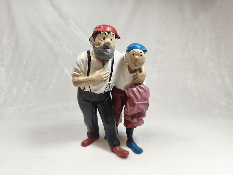 Op de afbeelding: Een geschilderd beeldje van twee figuren die samen staan. Het langere figuur heeft een grijze baard, een rode pet, een wit overhemd met zwarte bretels, een zwarte broek en rode schoenen. Het kleinere figuur draagt een blauwe pet en roze broek.
