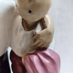 Op de afbeelding: Een geschilderd beeldje van een personage met een getinte huid, gekleed in een wit overhemd, een donkerrode broek en een roze kledingstuk dat over de arm is gedrapeerd. Het figuur heeft blauwe haardetails en een klein zwart puntje op de kin.