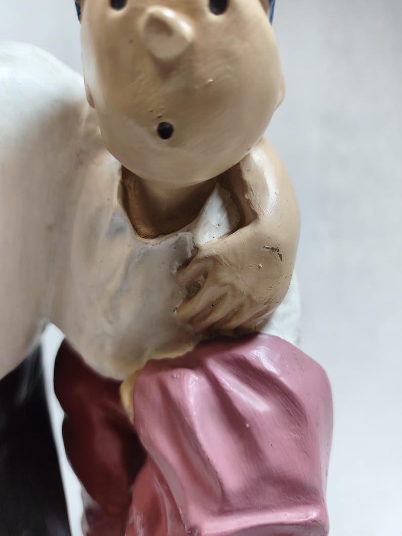 Op de afbeelding: Een geschilderd beeldje van een personage met een getinte huid, gekleed in een wit overhemd, een donkerrode broek en een roze kledingstuk dat over de arm is gedrapeerd. Het figuur heeft blauwe haardetails en een klein zwart puntje op de kin.